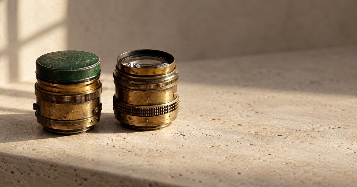 Two antique brass camera lenses side by side on pale limestone in warm side light, the left one capped with a deep-green lens cap.